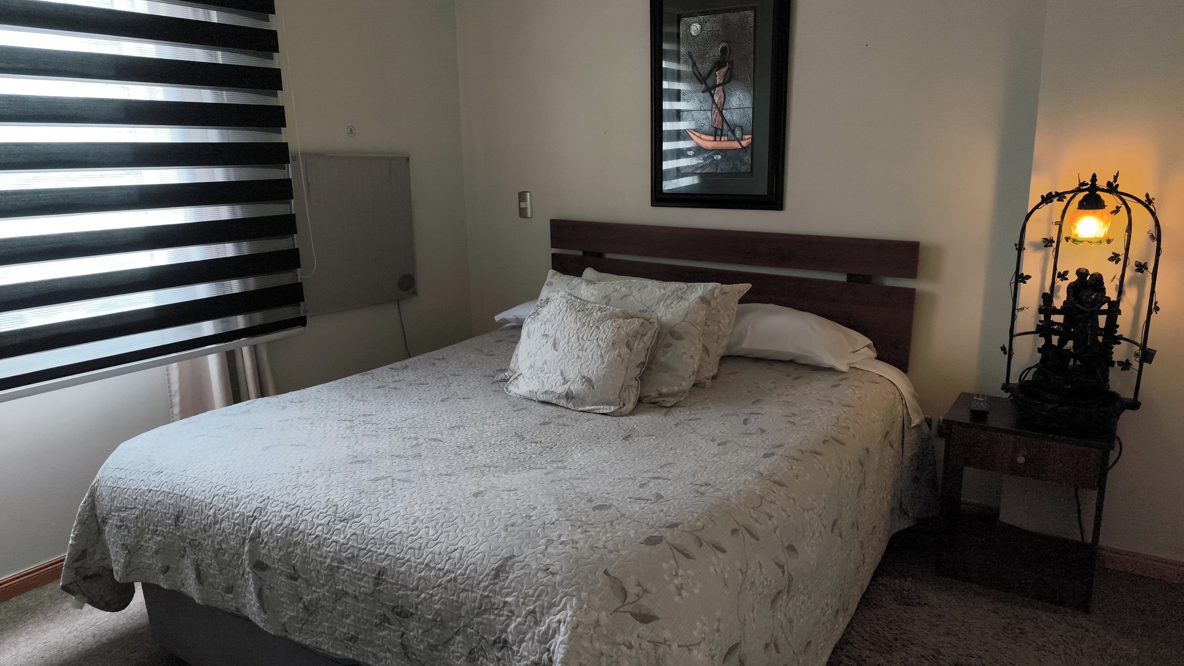 Uma cama bem arrumada com colcha estampada, cabeceira e mesa de cabeceira com abajur decorativo.