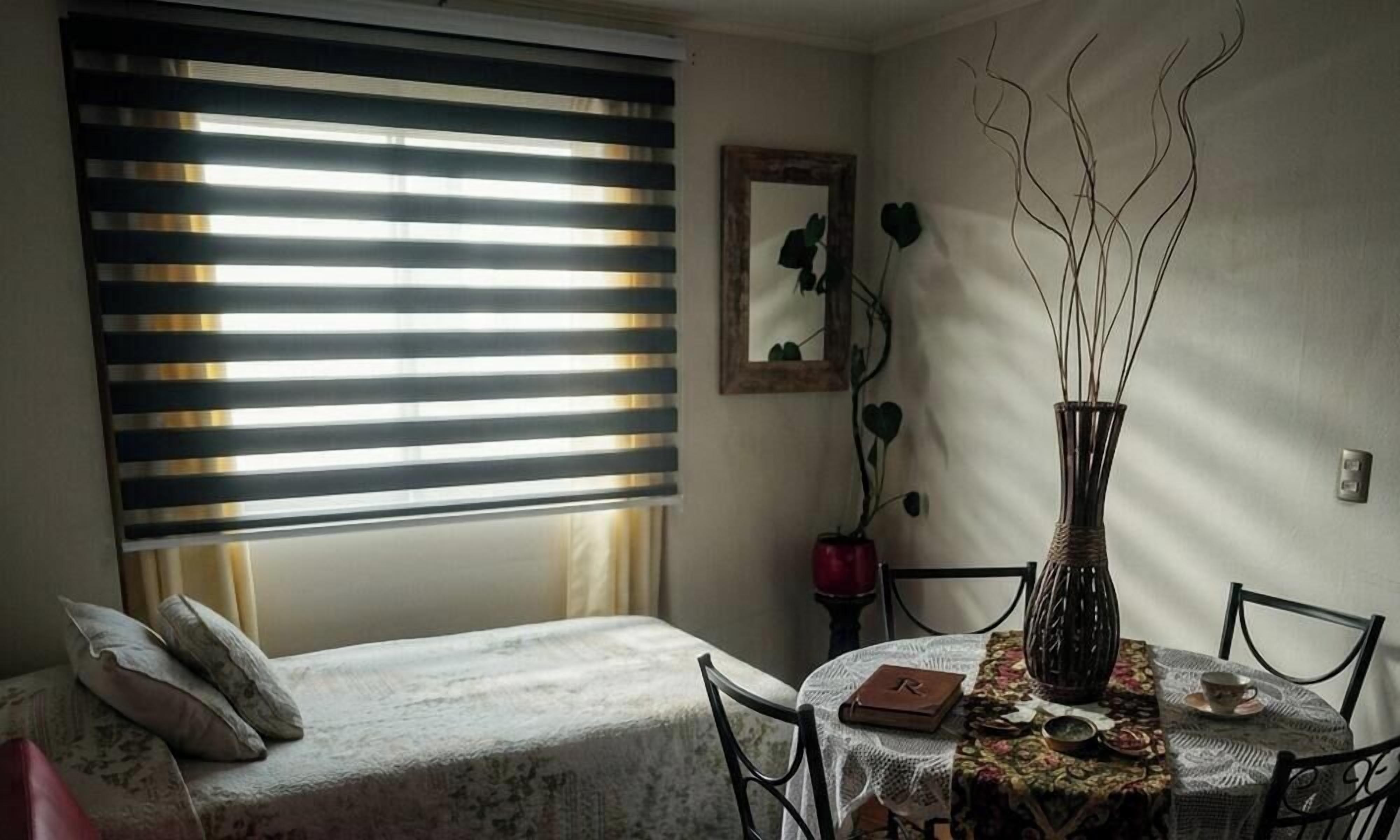 Una habitación pequeña con una cama, una mesa con un jarrón y una ventana con persianas.