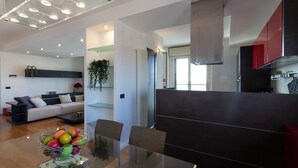 A modern kitchen with dark cabinetry, a dining area with a glass table, and a living room with a sofa and decorative plants. - notaMI - Settimo Cielo - San Cristoforo (Milan)