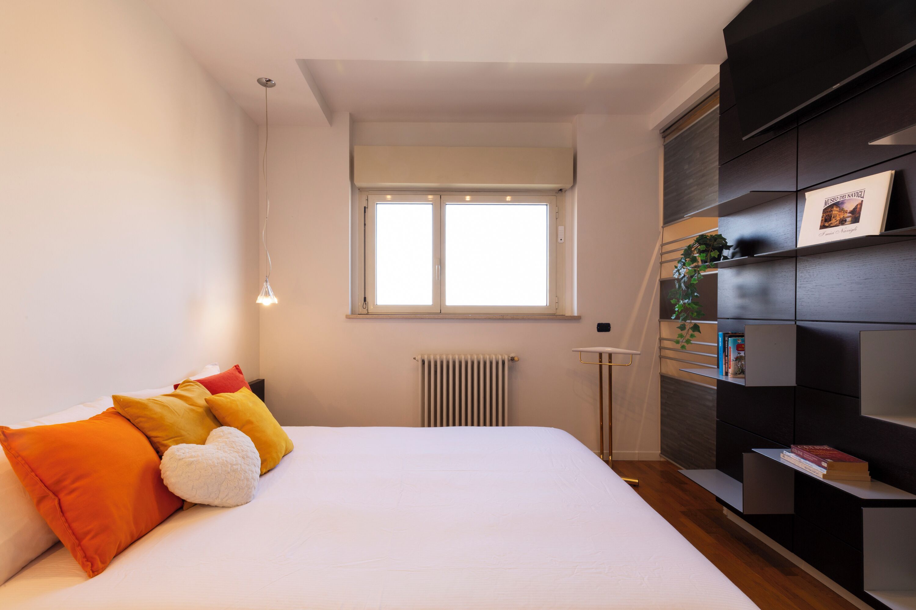 Een slaapkamer met een bed, kussens, een radiator, een raam en een boekenplank.