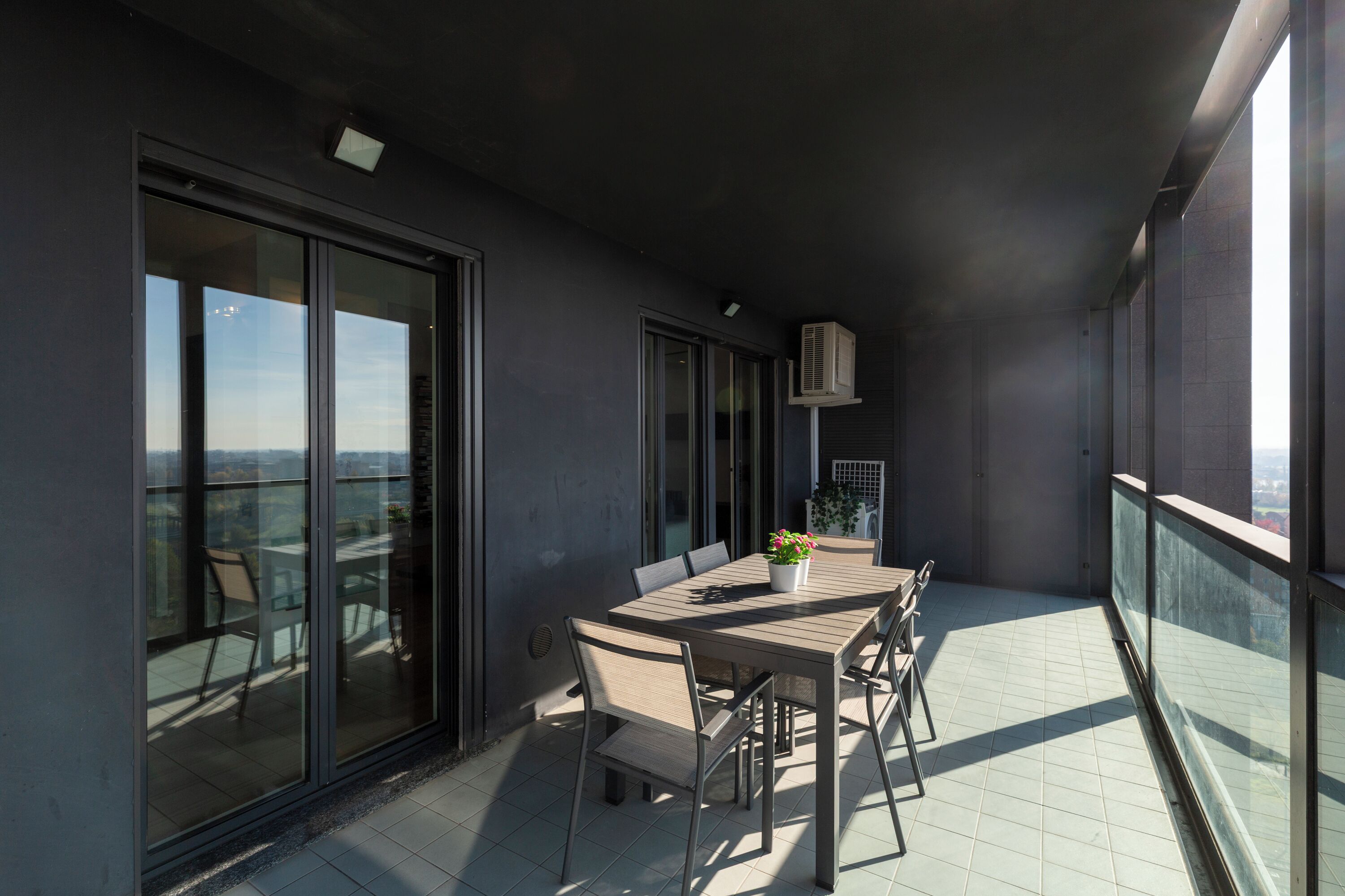 A balcony with a dining table and chairs, large windows, and an air conditioning unit.
