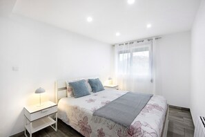 Un dormitorio con cama, dos mesitas de noche, lámpara, ventana con cortinas y un radiador.