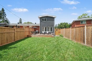 A backyard with a wooden deck, a small patio area, and a tall, narrow house. - Pet-Friendly 1BR With Office & Fenced Yard (Ottawa)