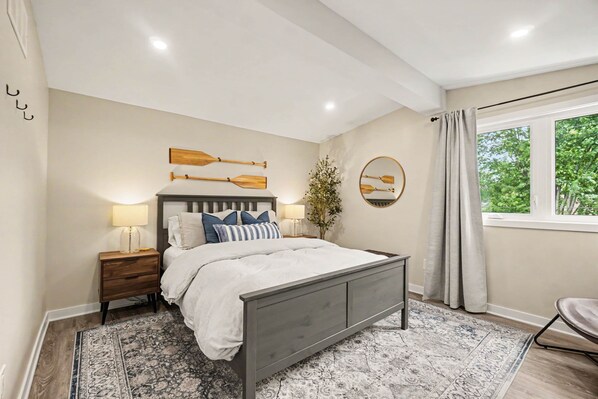A bedroom with a bed, nightstands, a wall-mounted mirror, and a window with curtains. - Pet-Friendly 1BR With Office & Fenced Yard (Ottawa)