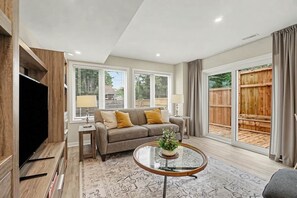 A living room with a sofa, a coffee table with a plant, a TV, and a large window with blinds. - Pet-Friendly 1BR With Office & Fenced Yard (Ottawa)