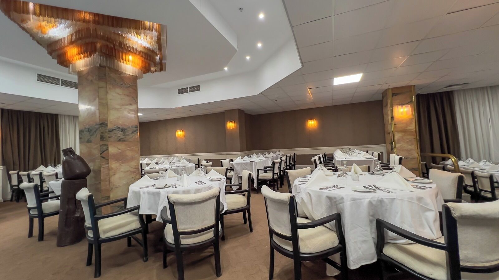 Une salle de banquet avec des tables rondes dressées pour le repas, une grande colonne décorative et des luminaires fixés au mur.