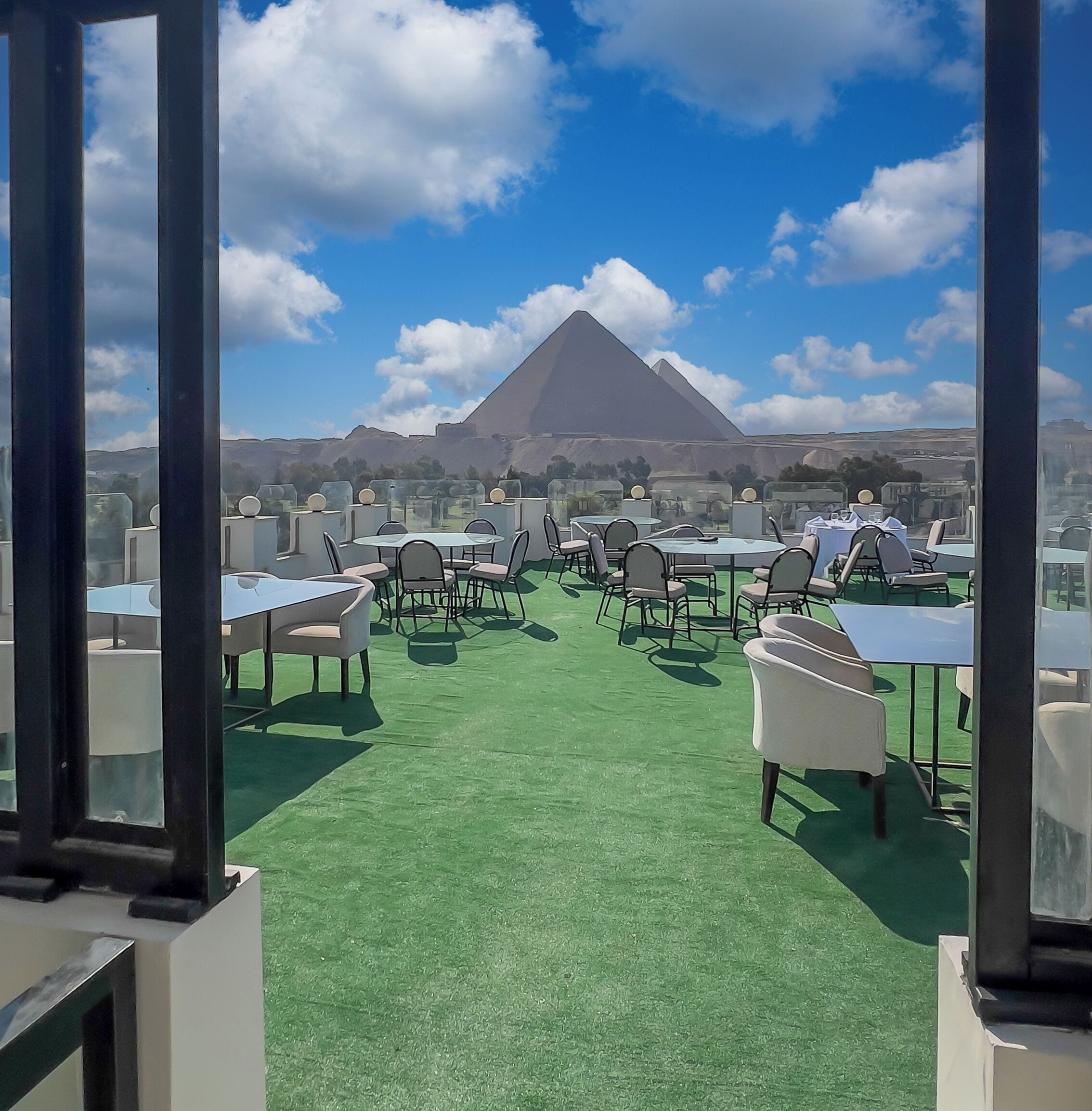 An outdoor dining area with tables and chairs, a green carpet, and a view of a pyramid in the background.
