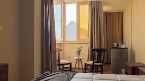 A hotel room with a bed, a small table with a vase, two chairs, a refrigerator, and a view of mountains through the window.