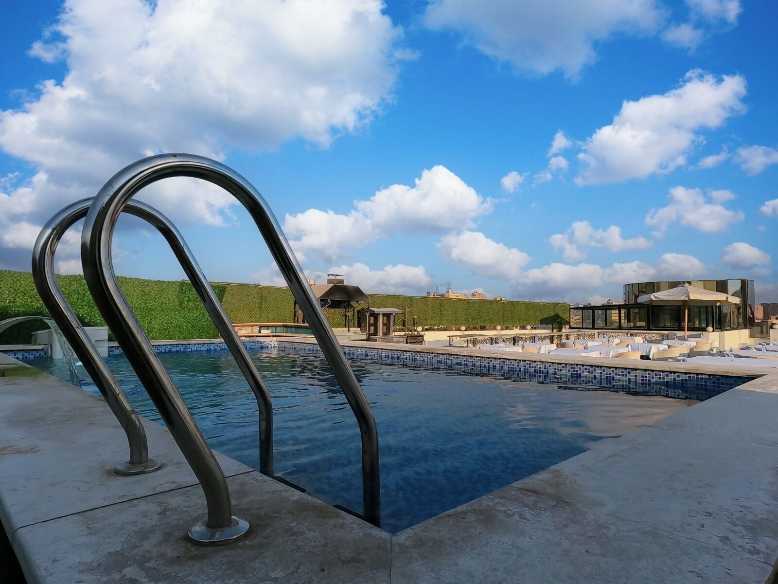 A swimming pool with metal railings, a green hillside, and a modern building with a glass facade in the background.