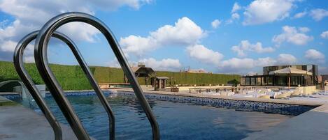 A swimming pool with metal railings, a green hillside, and a modern building with a glass facade in the background.