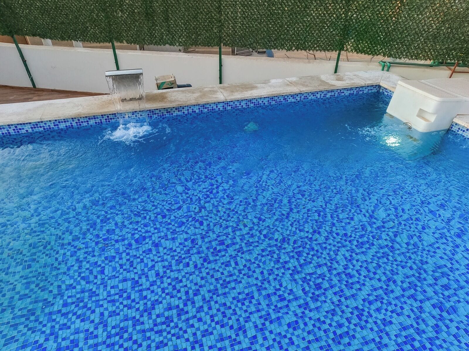 Una piscina con un diseño de mosaico azul, rodeada por un seto verde y una zona junto a la piscina de color blanco.