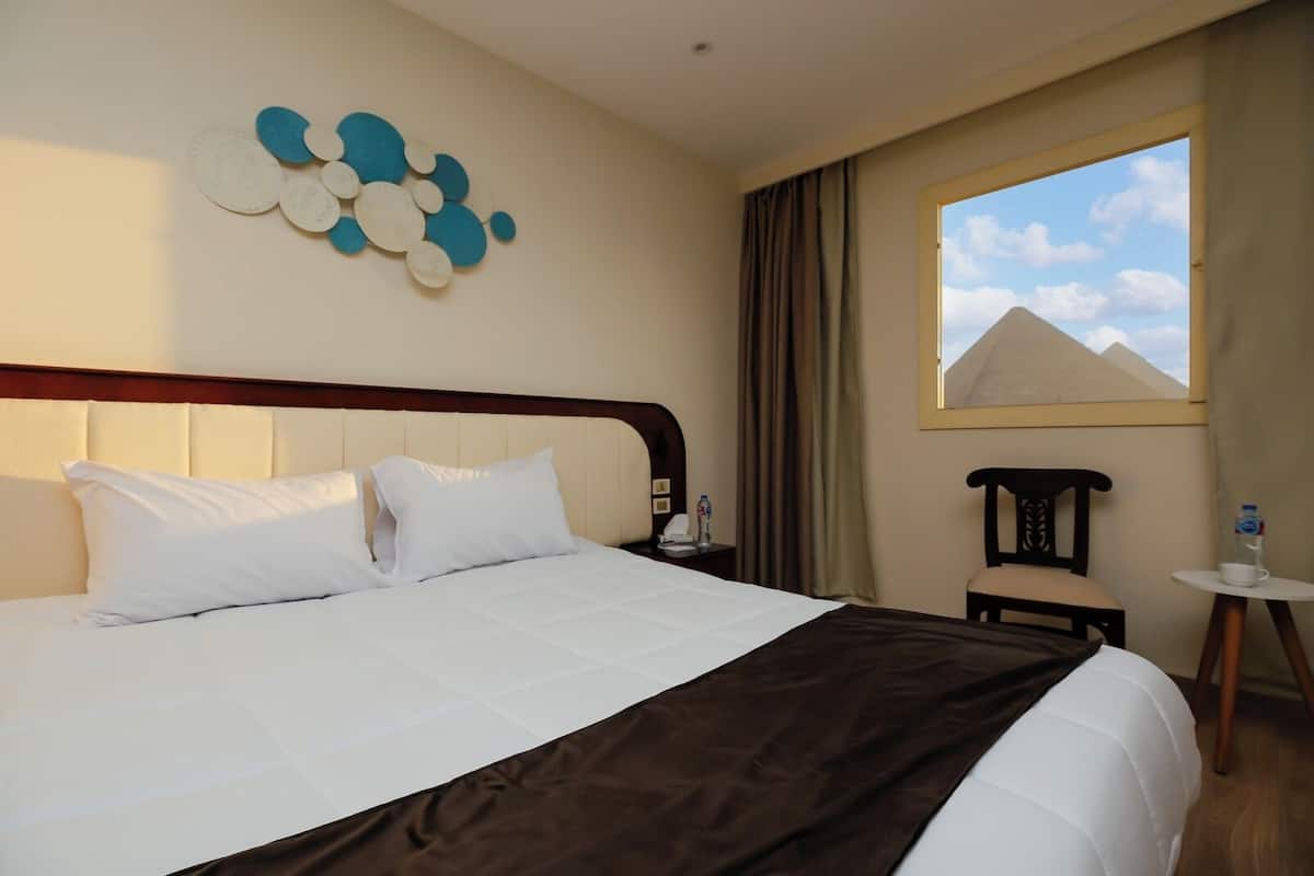 Une chambre d’hôtel avec un grand lit, une tête de lit, une table de chevet, une chaise, une petite table et une fenêtre donnant sur les pyramides.