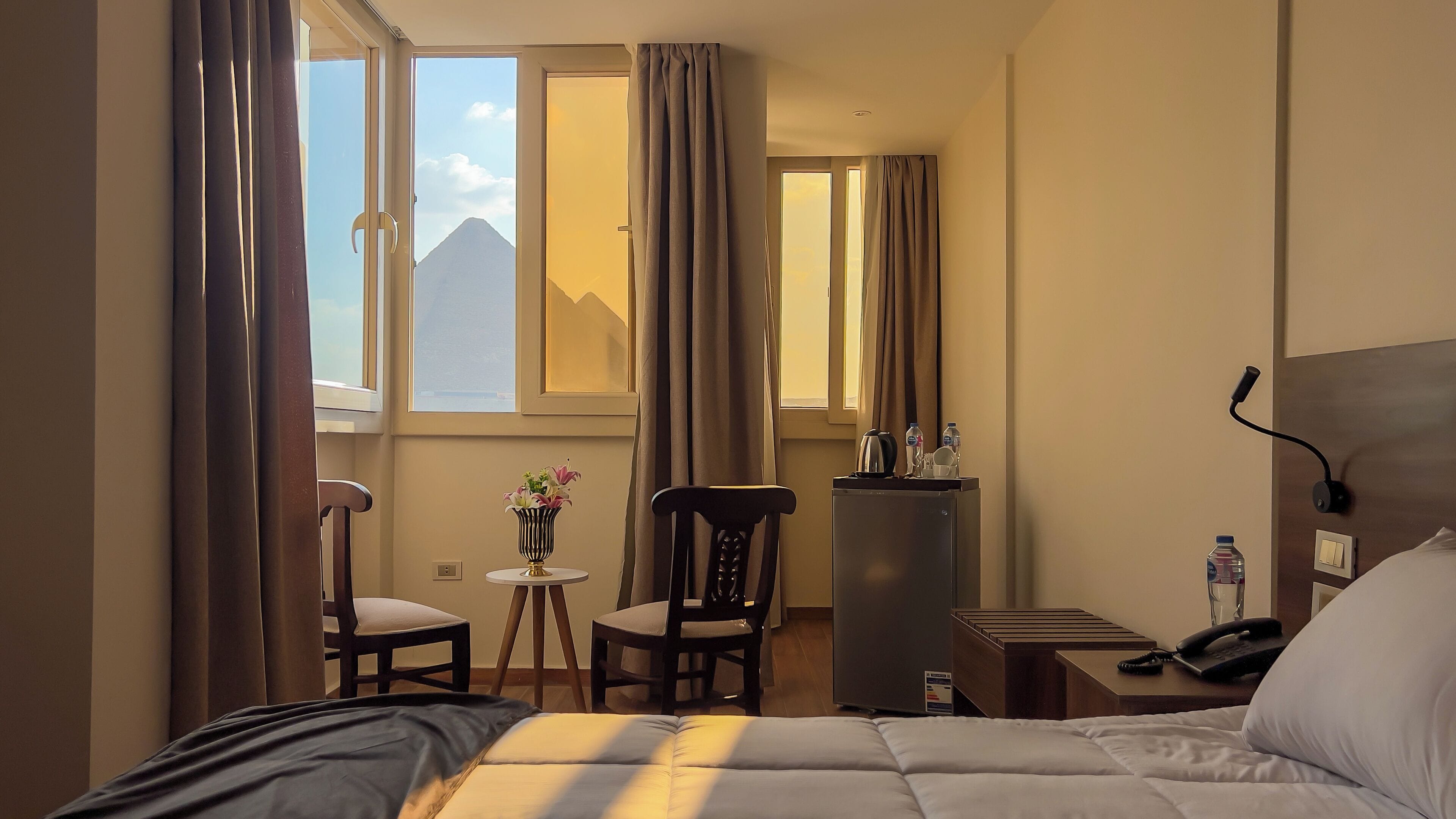 A hotel room with a bed, a desk, a chair, a small table with a vase, a refrigerator, a nightstand with a phone, and curtains.