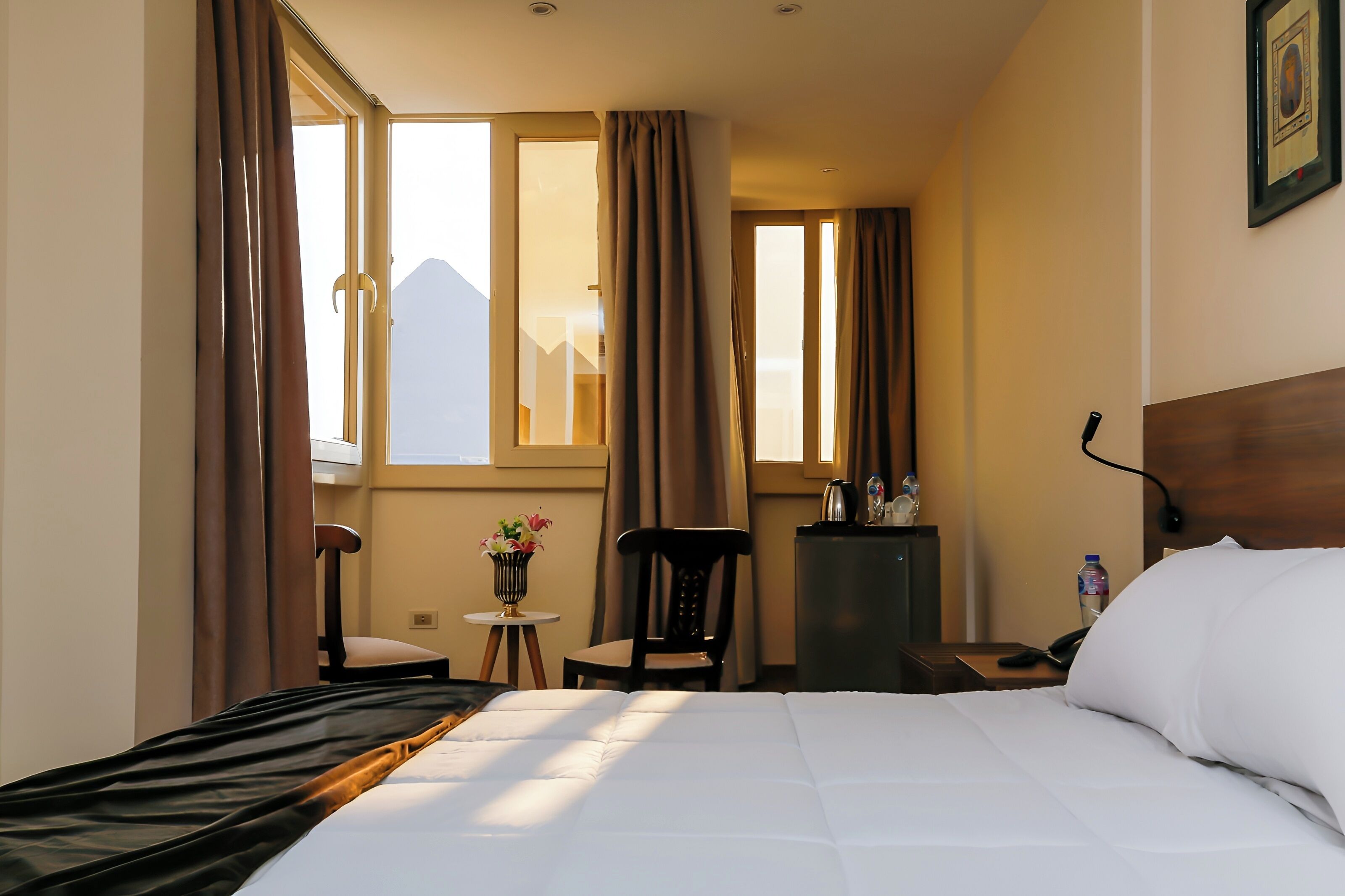 Habitación de hotel con una cama, una silla, una mesita con un florero, una mesita de noche con un teléfono y una ventana con cortinas.