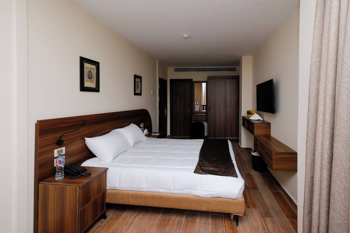 Une chambre d’hôtel avec un grand lit, une tête de lit en bois, une table de chevet avec un téléphone et une bouteille d’eau, un téléviseur à écran plat fixé au mur et un plancher en bois.