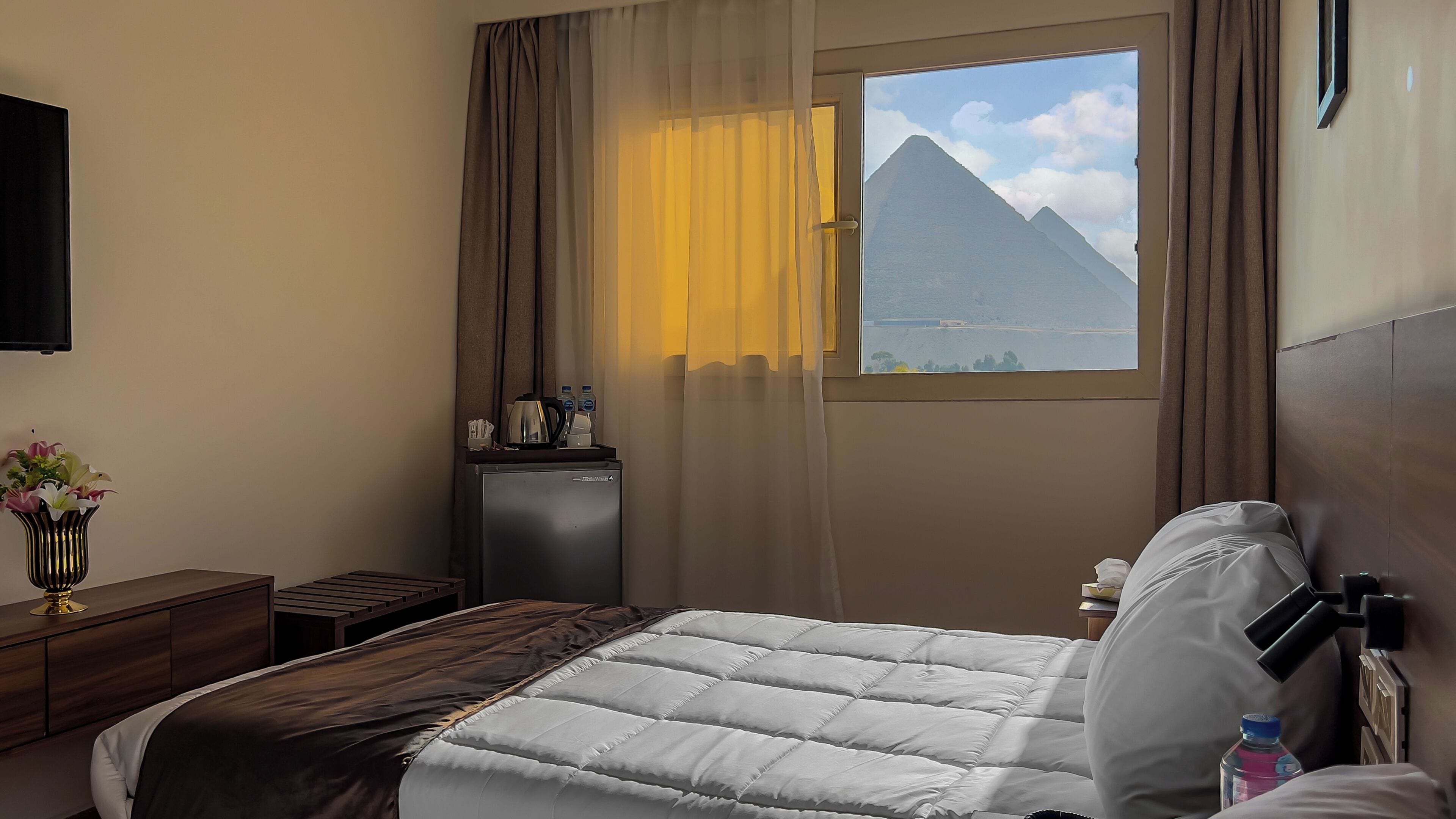 Habitación de hotel con cama, escritorio con cafetera, mesita de noche, televisor y ventana con vista a la montaña.