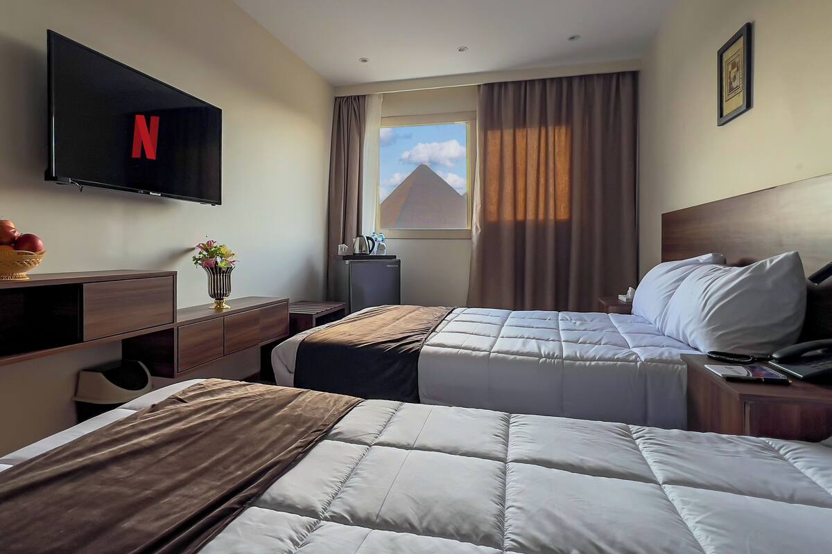 Une chambre d’hôtel avec un lit, un bureau, une télévision, une fenêtre avec des rideaux et une petite table sur laquelle se trouvent un panier de fruits et un vase.