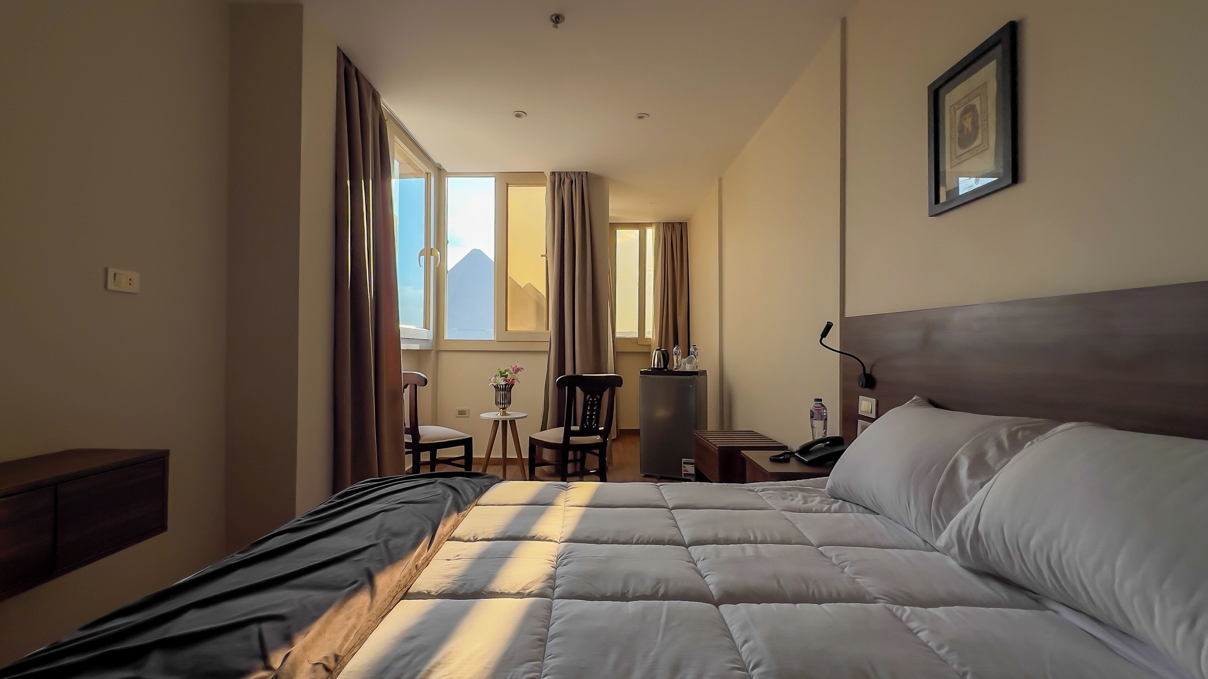 A hotel room with a large bed, a desk, a chair, a small table with a vase, and a view of the mountains through the window.