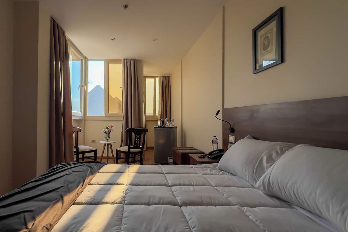 Une chambre d’hôtel avec un grand lit, un bureau, une chaise, une petite table avec un vase, et une vue sur les montagnes par la fenêtre.