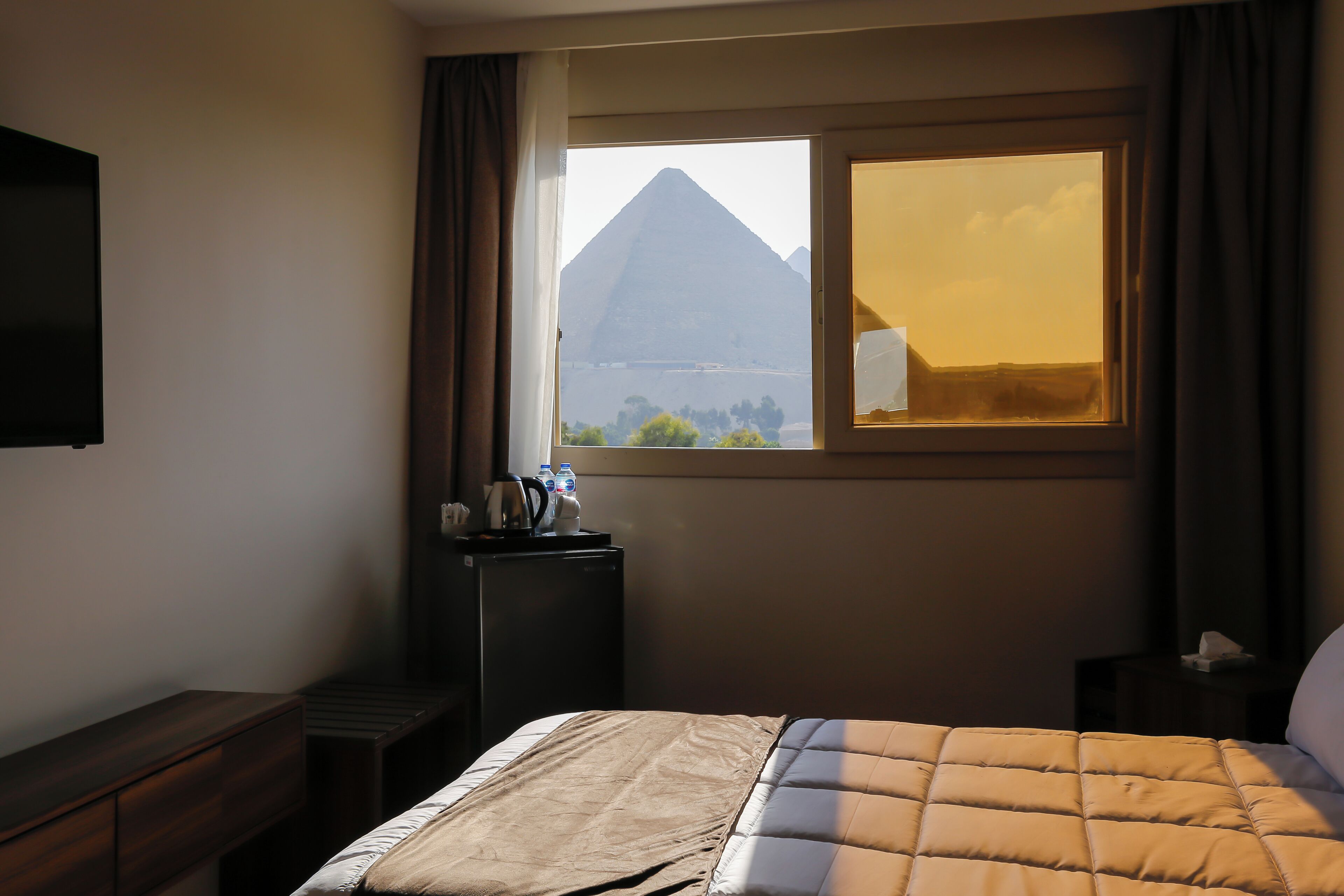A hotel room with a bed, a desk, a window with a view of a pyramid, and a flat-screen TV.