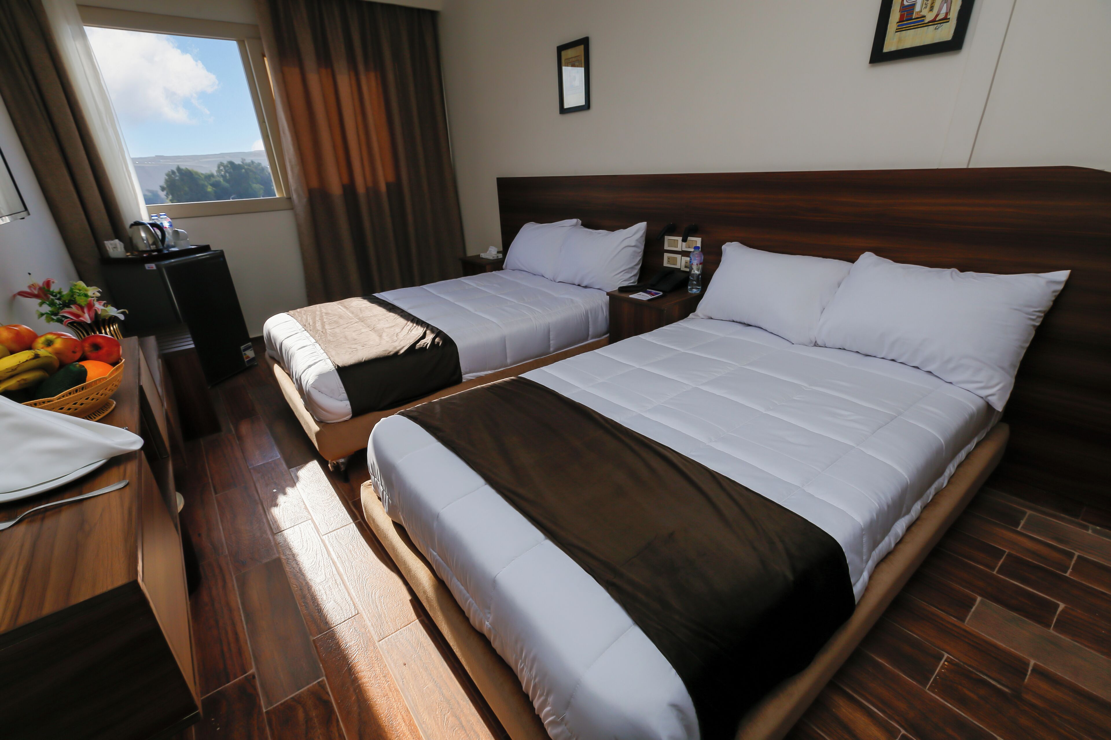 A hotel room with two beds, a wooden floor, a window with curtains, a desk with a basket of fruit, and a framed picture on the wall.