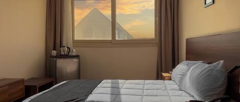 A hotel room with a bed, a nightstand, a desk, a window with a view of a mountain, and a small refrigerator.