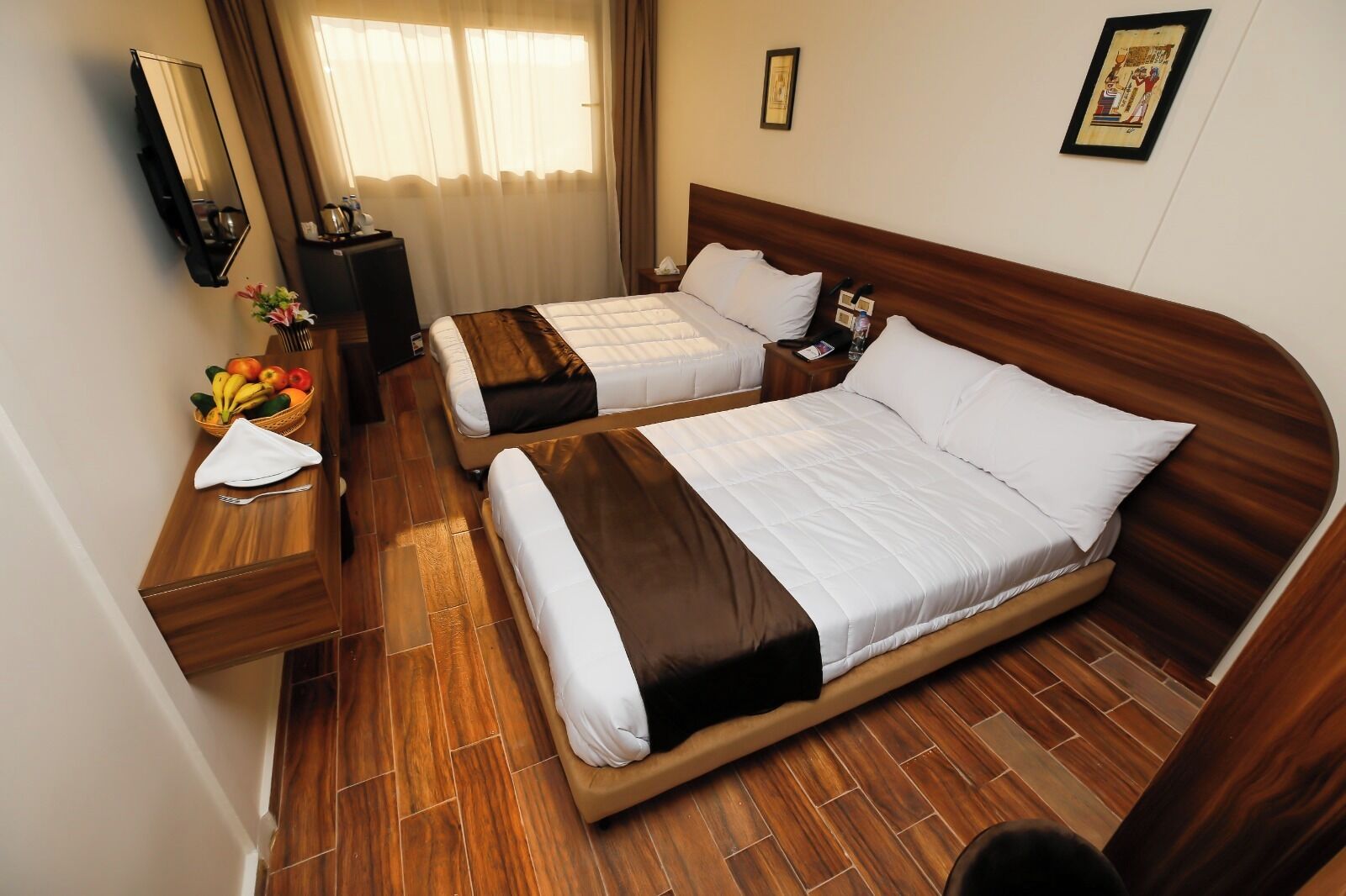 A hotel room with two beds, wooden flooring, a bedside table with a basket of fruit, a mirror, and a wall-mounted picture.