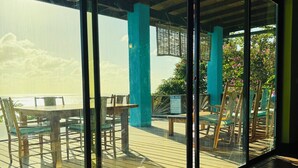 A covered outdoor seating area with tables and chairs, a wooden deck, and a view of the sea through large glass windows.