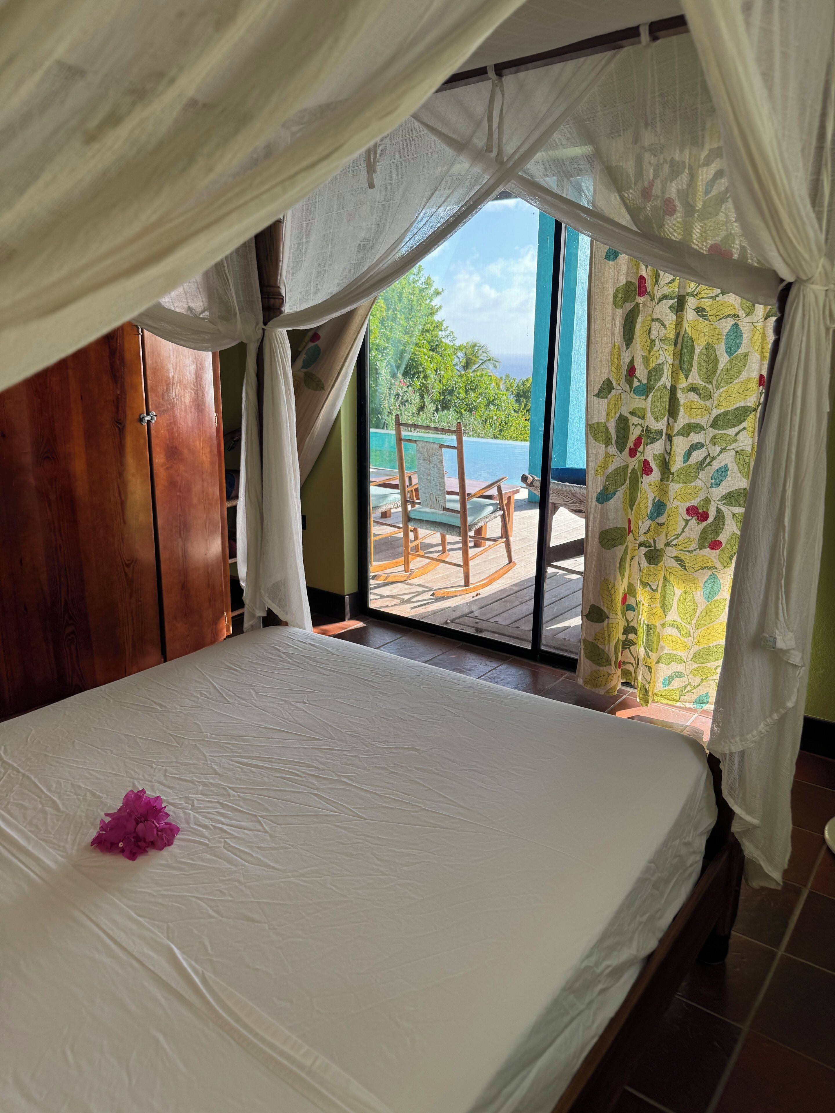 A bedroom with a bed, a wooden wardrobe, and a view of a pool and outdoor seating area.