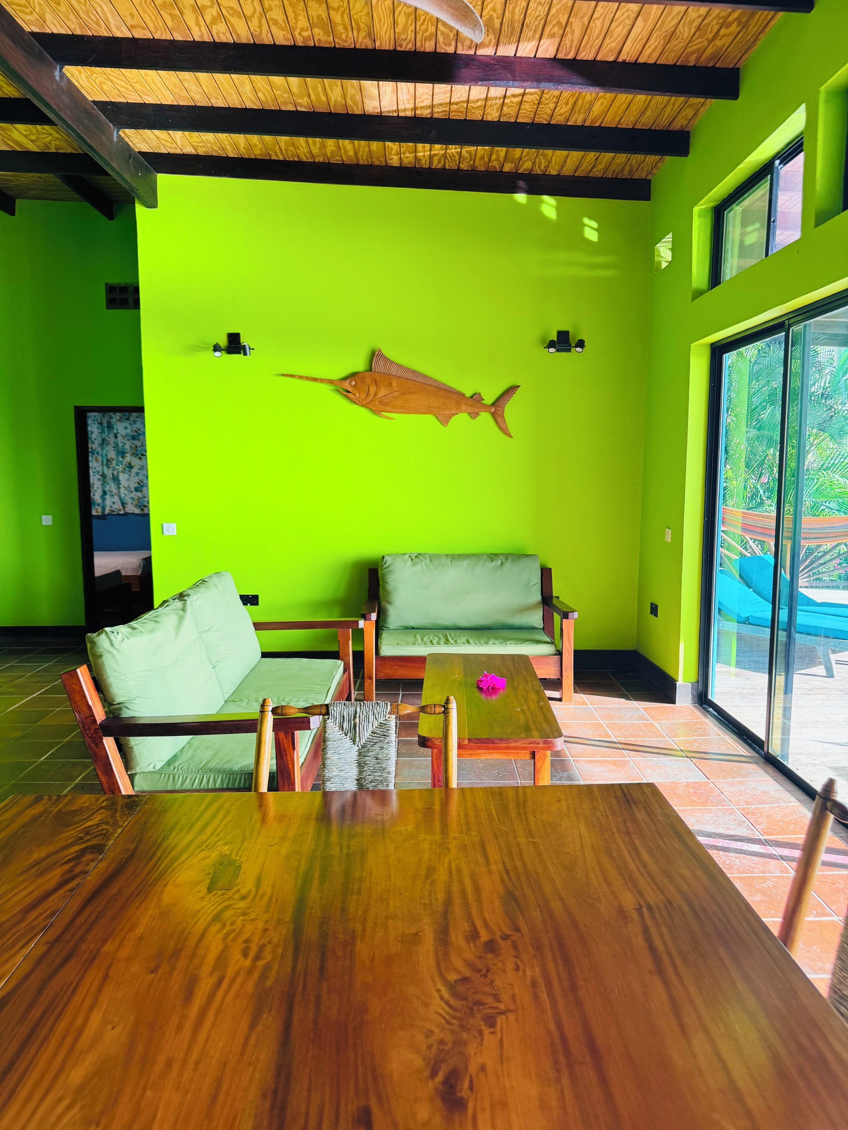 A room with a bright green wall, a wooden ceiling, and a large wooden table. There are two green cushioned chairs and a small wooden table with a pink flower.
