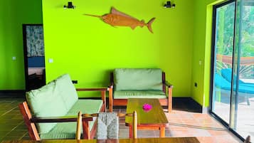 A room with a bright green wall, a wooden ceiling, and a large wooden table. There are two green cushioned chairs and a small wooden table with a pink flower.