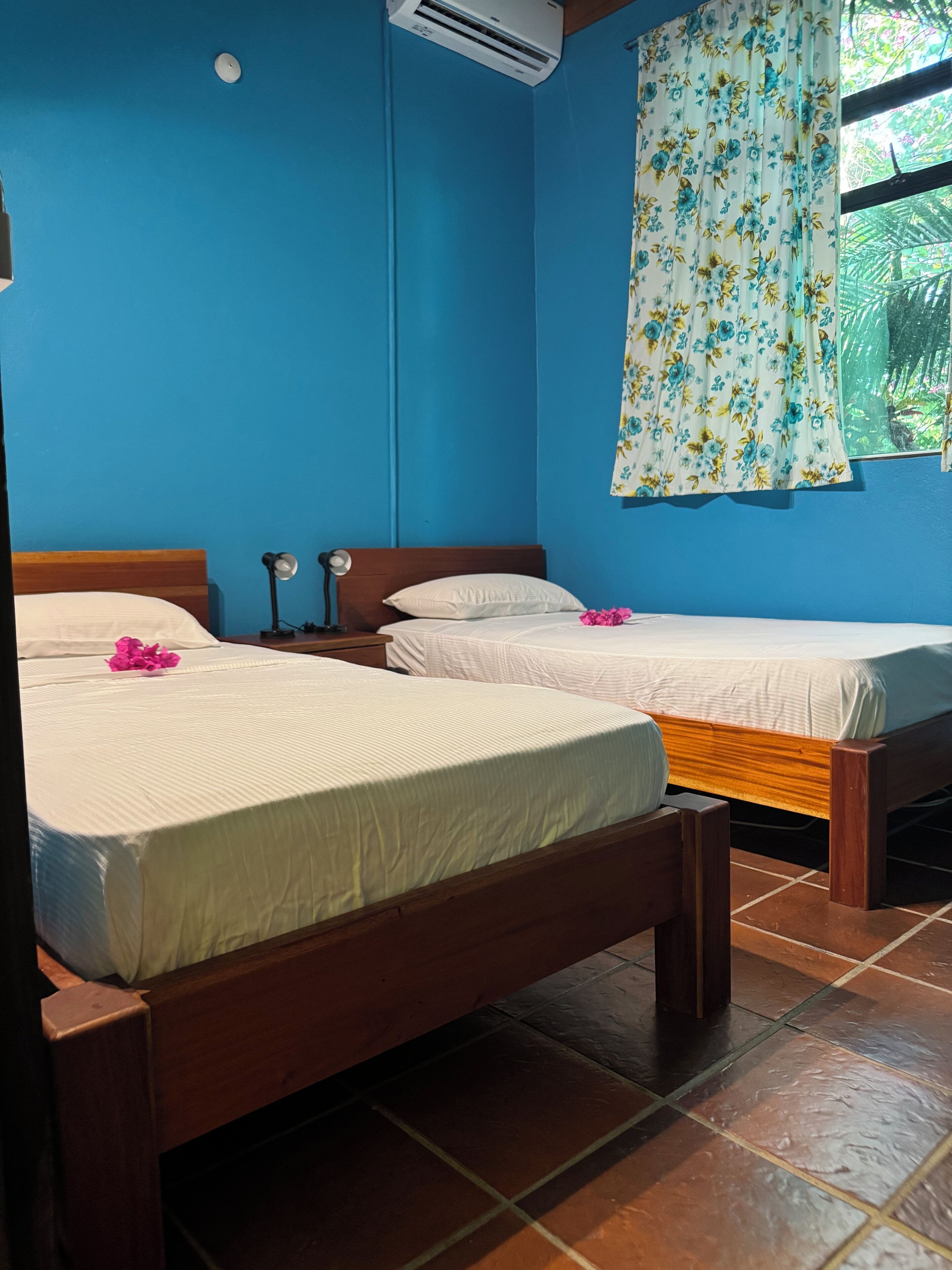 A room with two wooden beds, a blue wall, a window with floral curtains, and a tiled floor.