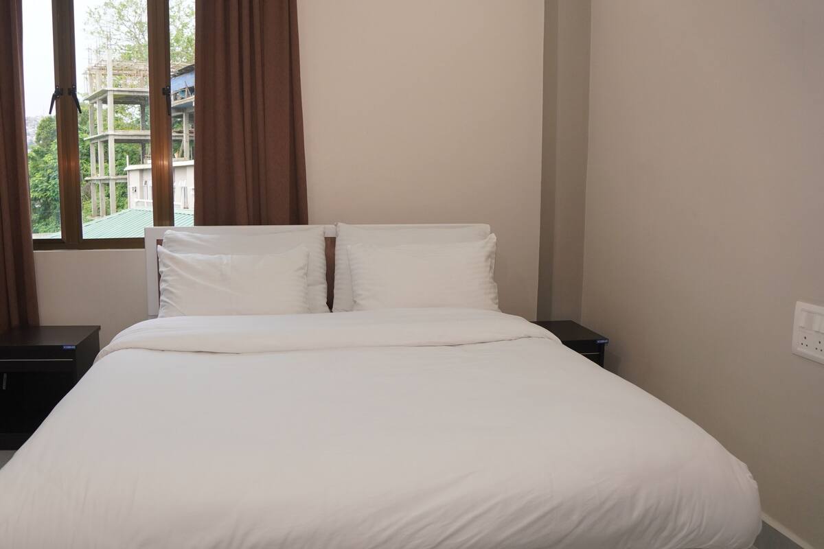 A bedroom with a bed, two bedside tables, a window with curtains, and a view of a building outside.