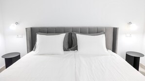 Une chambre à coucher moderne avec une tête de lit rembourrée de couleur grise, une literie blanche et deux tables de chevet avec des lampes.