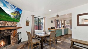 A dining area with a stone fireplace, wooden table, and chairs. A kitchen is visible in the background with a window and a view of the outdoors. - Slopeside Cabin C W Hot Tub & Slope View Parking (Big Bear Lake)