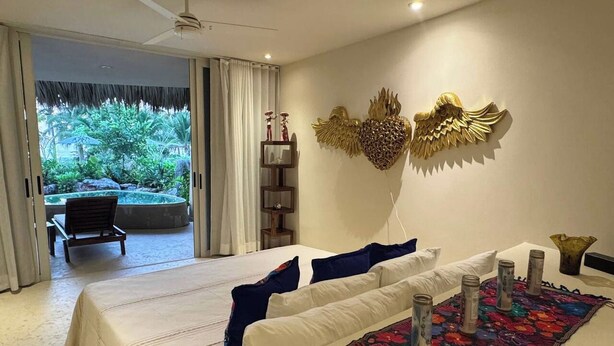 Een slaapkamer met een bed, kussens, een decoratief kunstwerk en uitzicht op een buitenruimte met een stoel en planten.