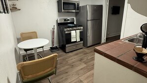A compact kitchen with white walls, a wooden floor, a white dining table with two chairs, a microwave, a stove, and a refrigerator. - 1BR Minutes to Stockyards • Fast Wi-Fi + Parking (Fort Worth)