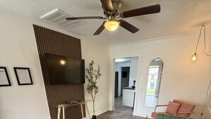 A modern living room with a green sofa, wooden flooring, a ceiling fan, a TV, and a small plant. - 1BR Minutes to Stockyards • Fast Wi-Fi + Parking (Fort Worth)