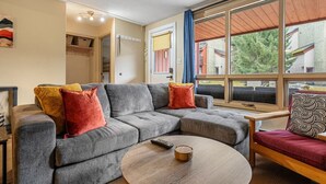 A modern living room with a grey sectional sofa, a round wooden coffee table, and a red armchair. A large window offers a view of the outside. - The Keep | Ski-In/Out Toby Chair | Ground Floor (Panorama)
