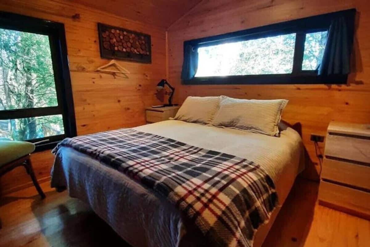 A wooden cabin bedroom with a bed, a nightstand, a chair, and a window.