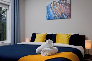 A hotel room with a bed, pillows, a towel, a painting on the wall, and a window with curtains. - Downtown Golden Serenity Suite – Riverstone Condo (Golden)