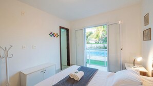 A hotel room with a bed, a bedside table with a lamp, a wardrobe, a mirror, and a view of a pool and greenery outside. - House w/ Pool 200 m From the Beach Pro0003 (Camaçari)