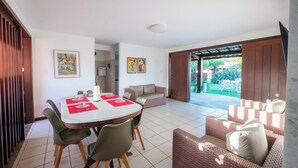A dining area with a round table and chairs, a sofa, and a view of a garden through an open door. - House w/ Pool 200 m From the Beach Pro0003 (Camaçari)