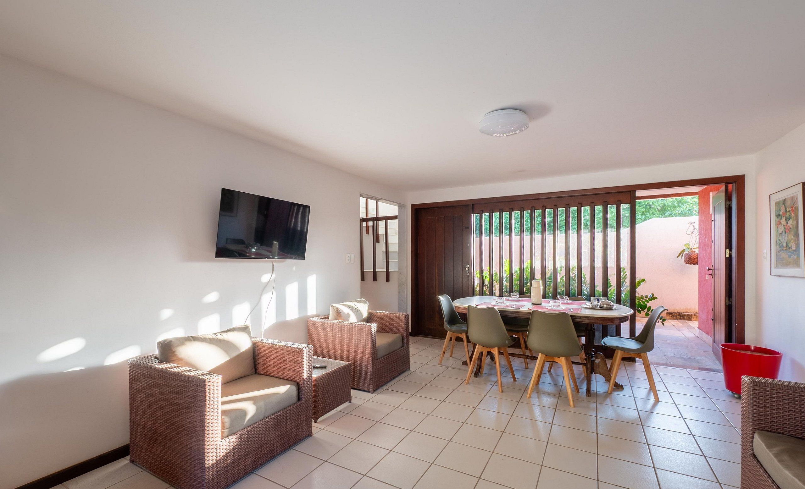 Ampla sala de estar com uma mesa de jantar posta para quatro pessoas, rodeada por móveis de vime. Uma televisão de tela plana está fixada na parede, e uma porta de correr dá acesso a uma área externa.