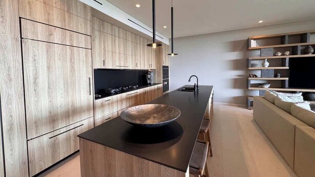 A modern kitchen with wooden cabinetry, a central island with a bowl, pendant lighting, and a built-in TV shelf. - Lujo Frente al Mar para 8 (Nay.)