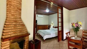 A room with a brick fireplace, a bed with a white bedspread, a wooden chair, and a table with a vase of flowers. - Pousada Caminho das Candeias (Nova Friburgo)