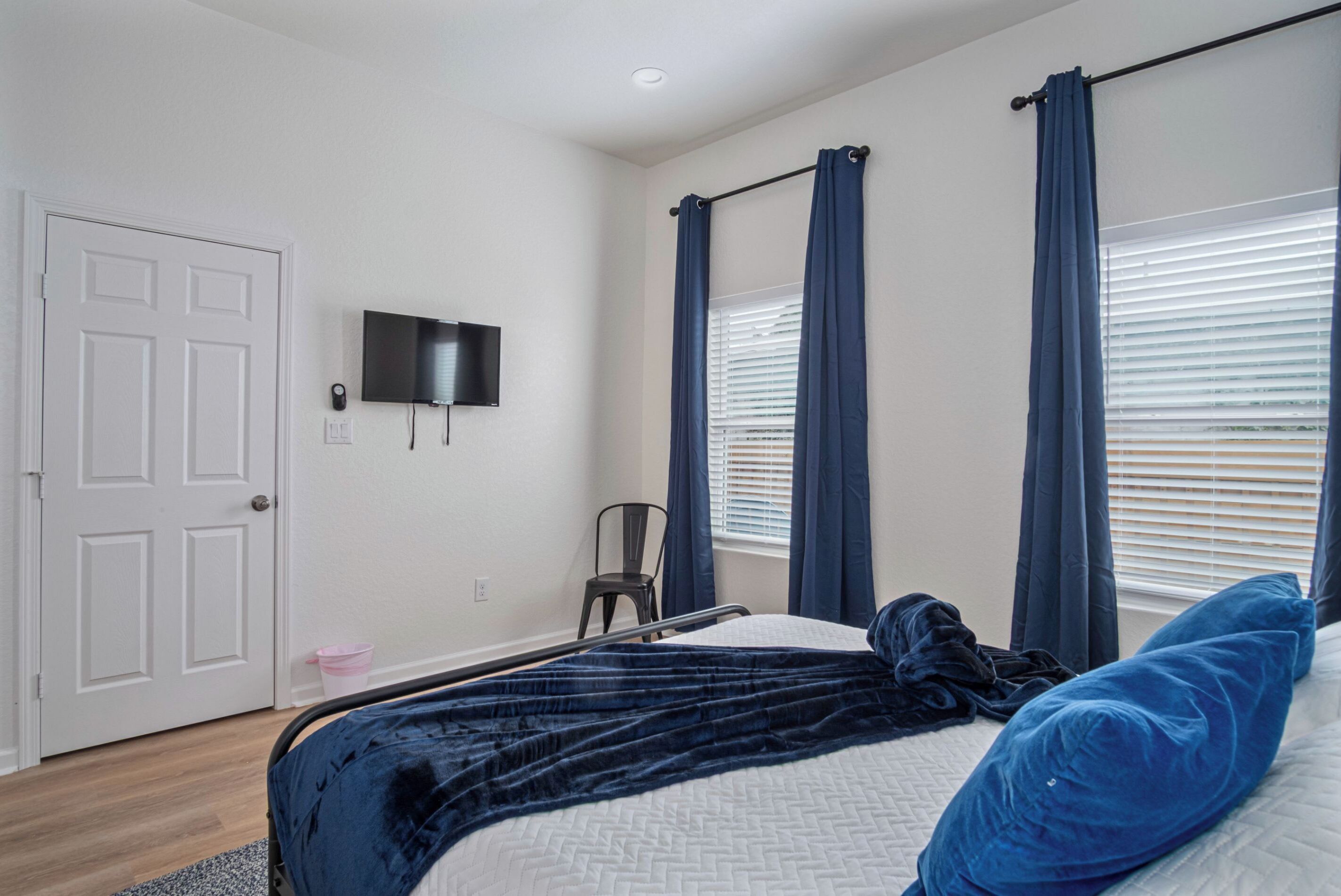 Une chambre à coucher avec un lit, un téléviseur fixé au mur, une porte et une fenêtre avec des rideaux.