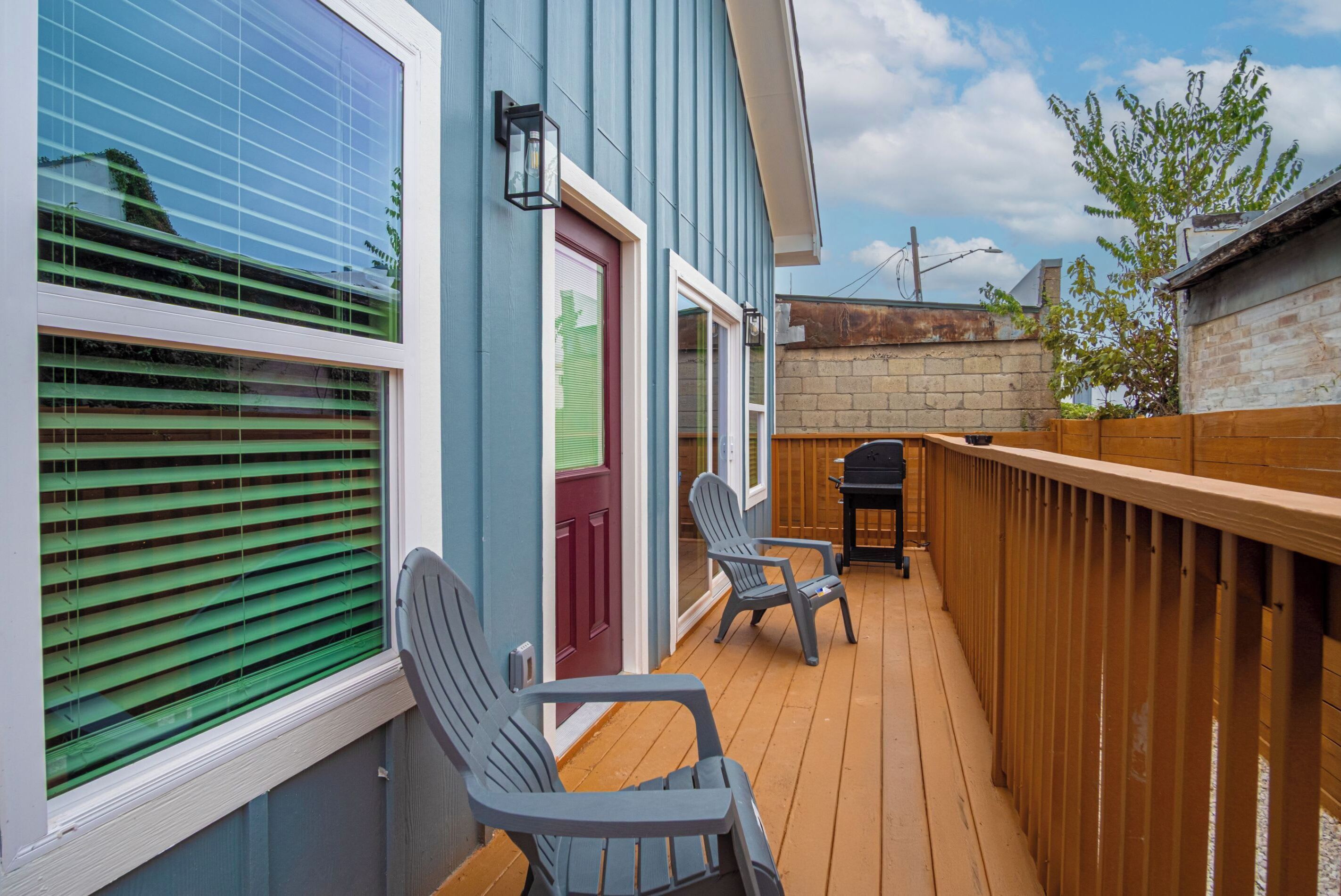 A wooden deck with two Adirondack chairs, a red door, and a brick wall.