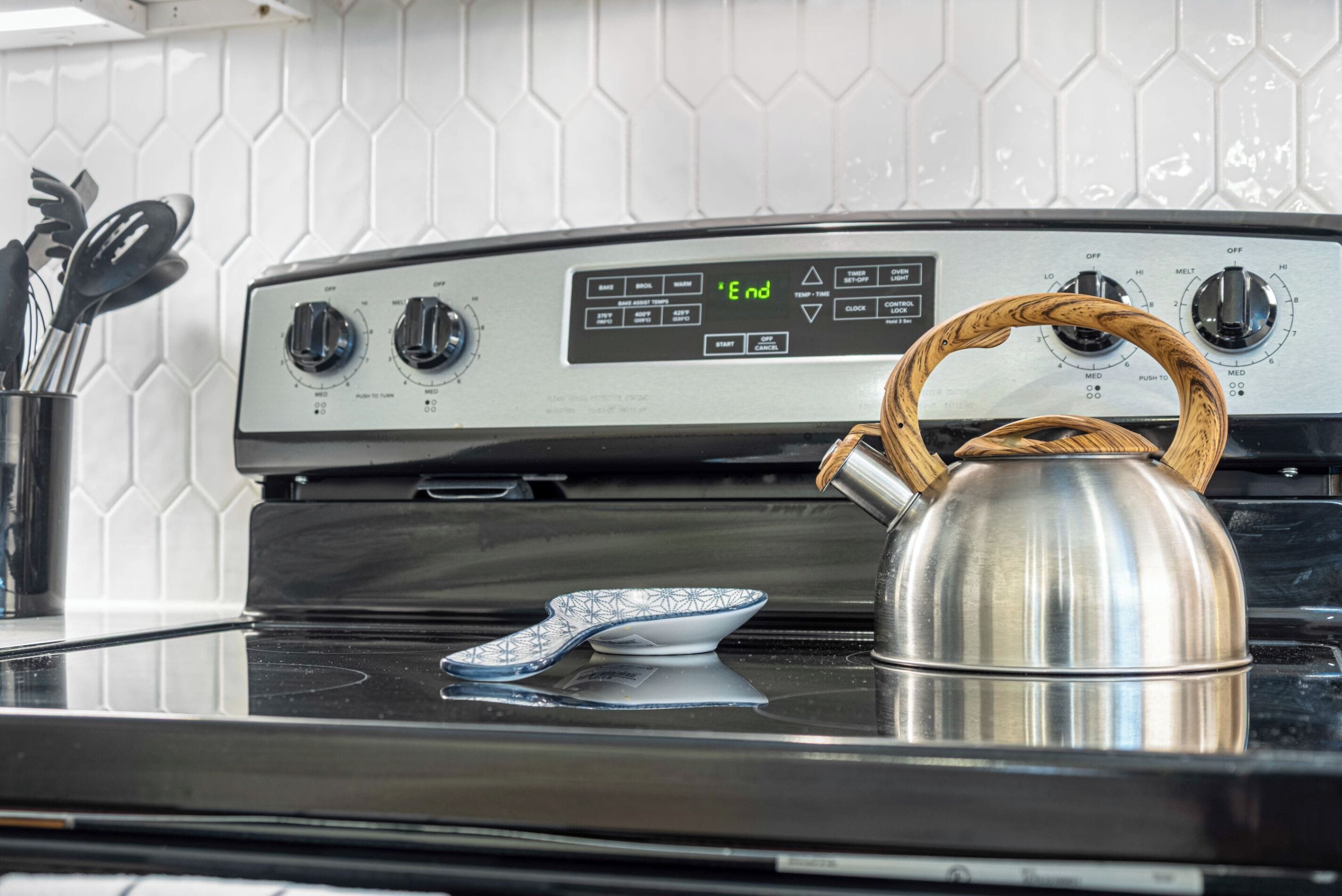 Une cuisine avec une bouilloire en acier inoxydable sur la cuisinière, une horloge numérique affichant le mot « Fin », et un pare-feu en carreaux hexagonaux blancs.
