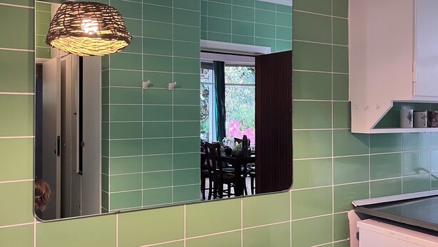 Une pièce aux murs recouverts de carreaux verts, avec une table en bois, un luminaire suspendu en osier et un miroir qui reflète un coin repas.